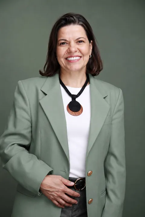 Retrato da Dra. Daniela Gomes, médica nutróloga pediátrica, sorrindo, vestindo blazer verde-sálvia, diante de um fundo verde-oliva.